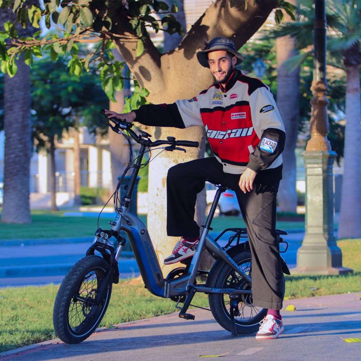 Persona seduta su una bicicletta elettrica. L'uomo indossa una giacca e un cappello. La bicicletta è grigia e blu. Lo sfondo è una strada.