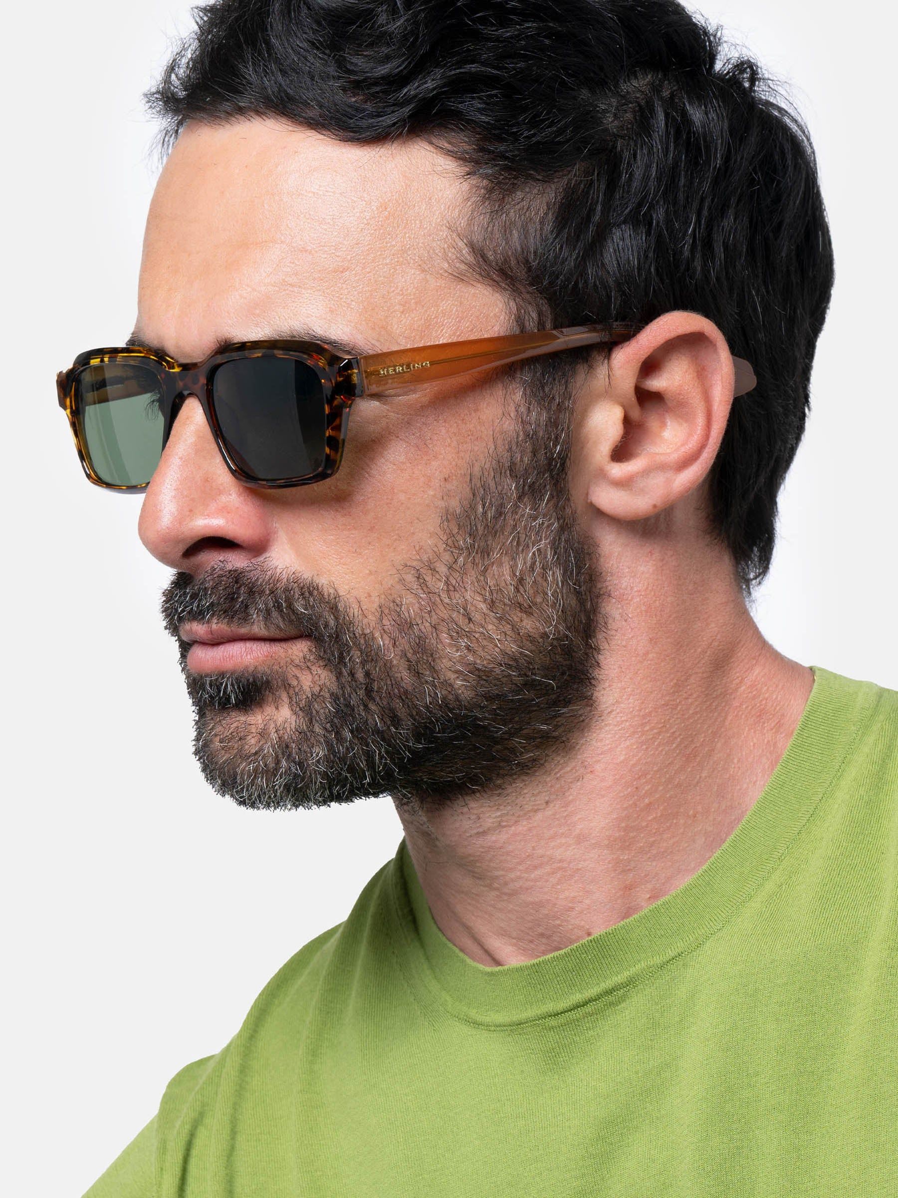 Uomo con occhiali da sole. Gli occhiali hanno una montatura marrone e lenti verdi. L'uomo indossa una maglietta verde.