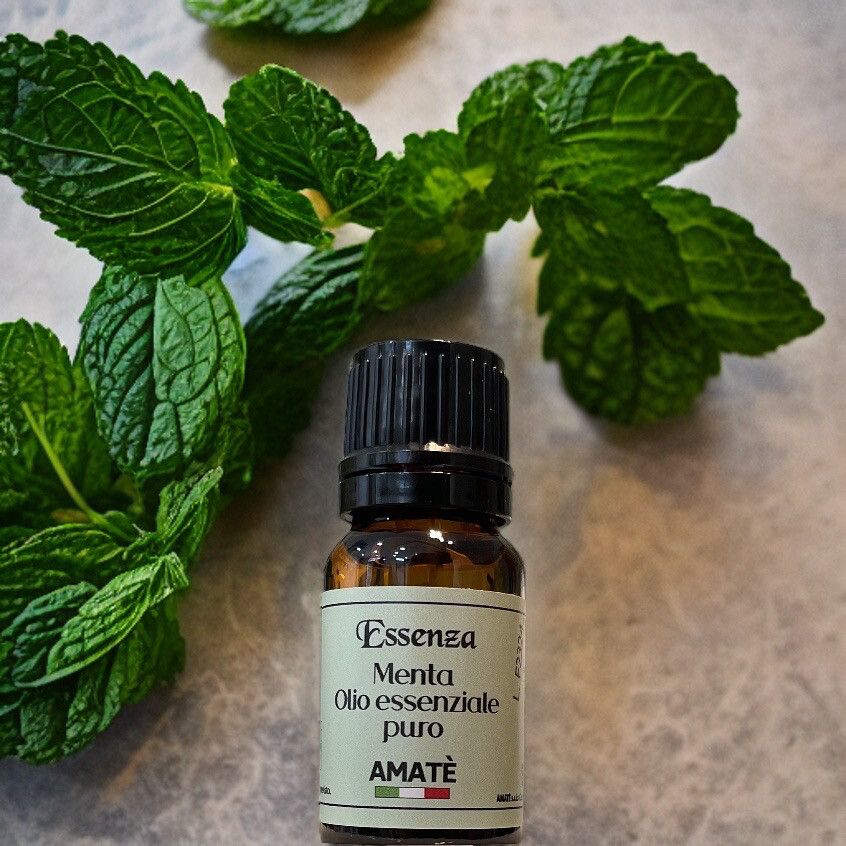 Flacone di vetro marrone con tappo nero, circondato da foglie di menta. Etichetta: Essenza Menta Olio essenziale puro. Marchio: AMATÈ.