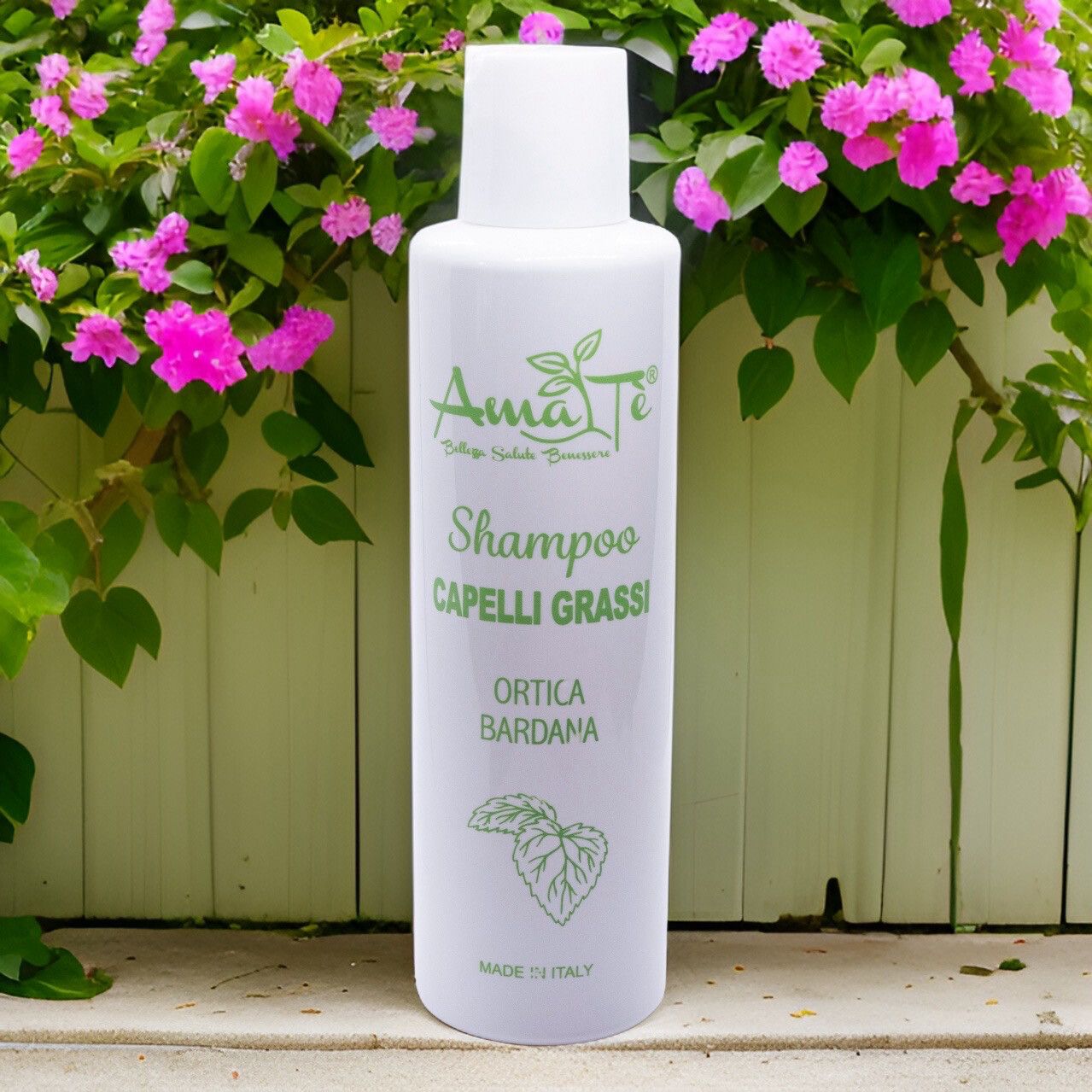 Flacone bianco con etichetta verde. Testo: Amatè Shampoo Capelli Grassi Ortica Bardana. Sfondo: fiori.