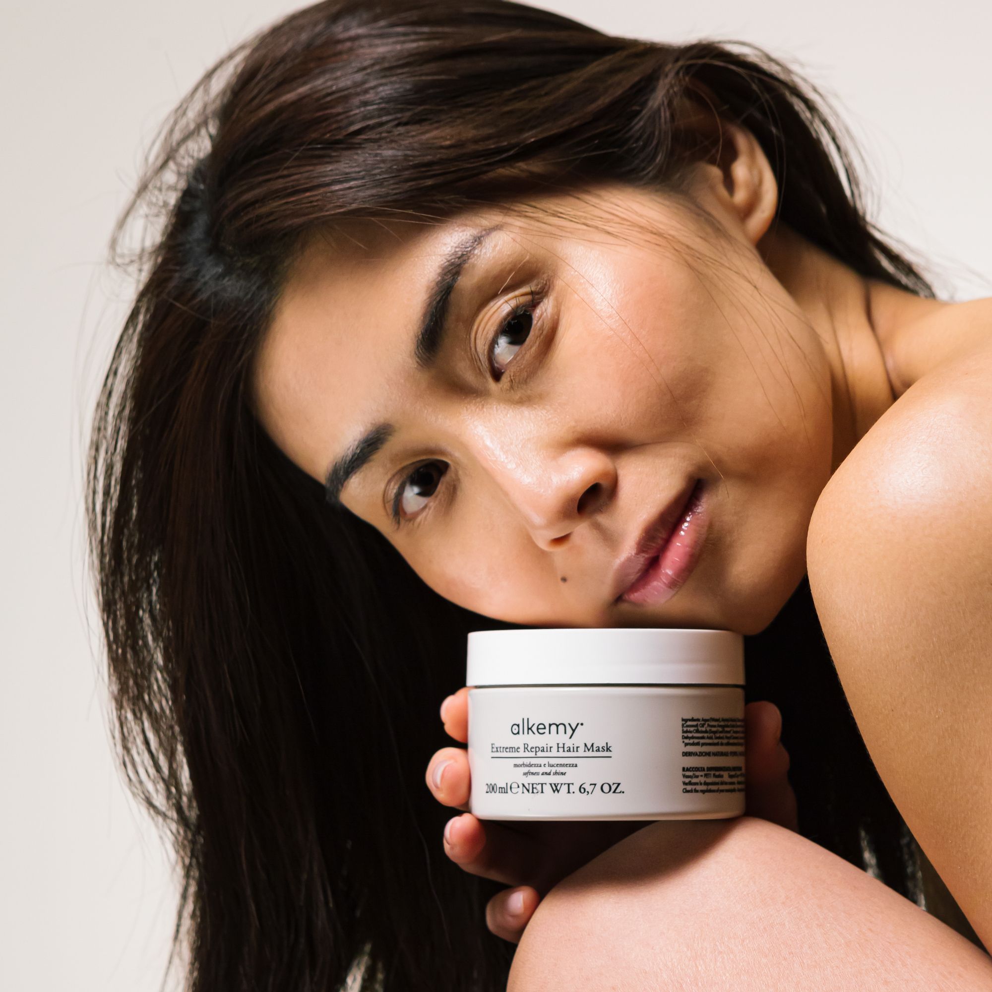 Alkemy Maschera Ristrutturante Intensa in un barattolo color crema, tenuto da una donna.