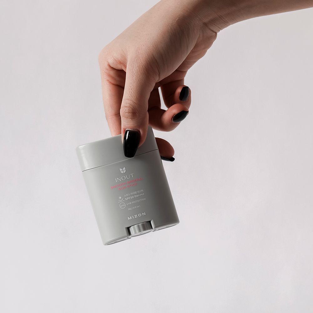 Mano che tiene uno stick solare grigio. Testo: INOUT, Smooth Mineral Sun Stick, SPF 50 PA++++, MIZON. Unghie nere.