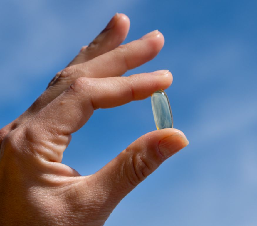 Mano che tiene una capsula molle di olio di pesce Omega-3 Aavalabs davanti al cielo blu.