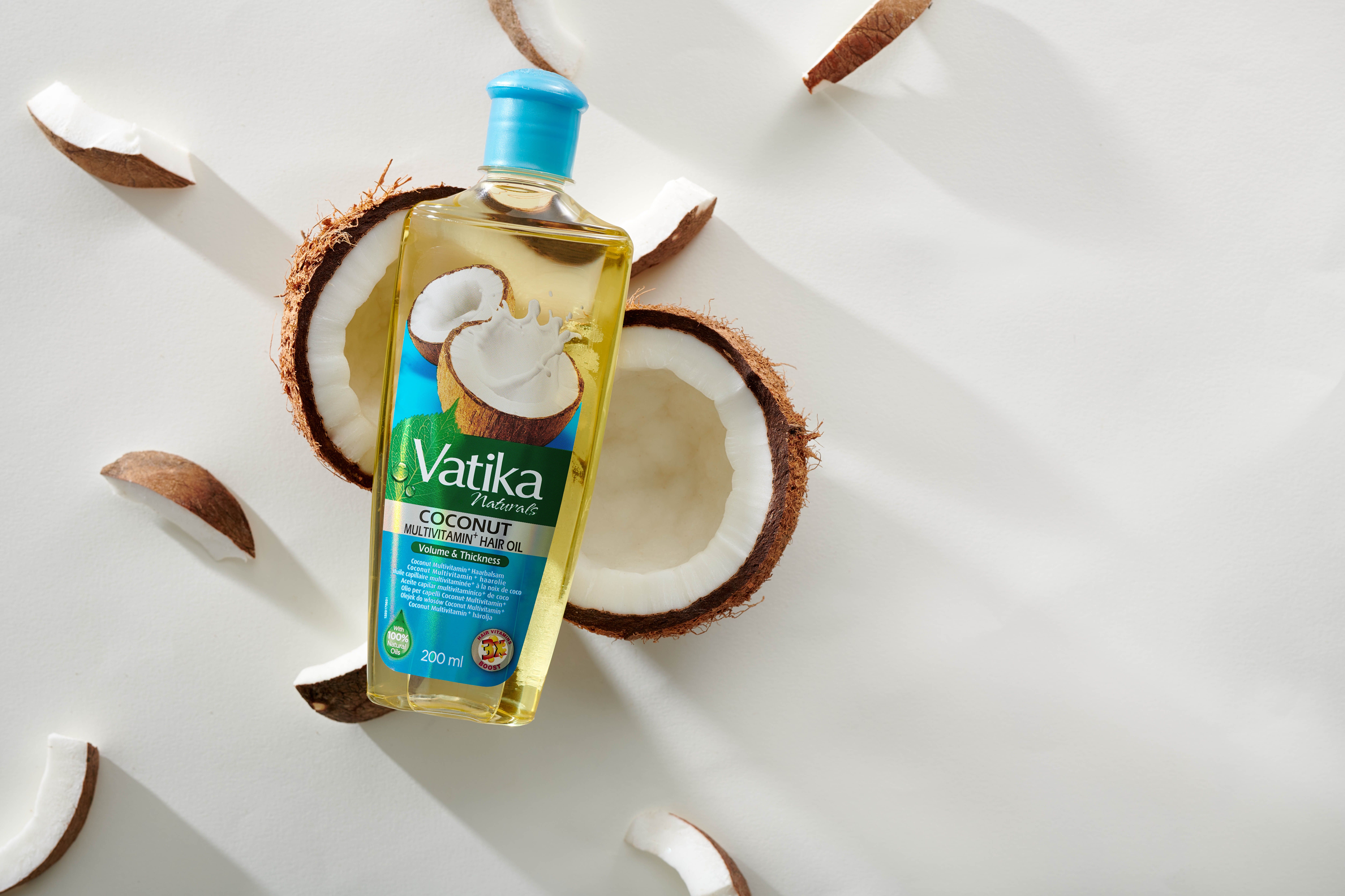 Flacone di olio per capelli Vatika Naturals, tappo blu, circondato da metà e pezzi di cocco. Scritta: Coconut Multivitamin Hair Oil.