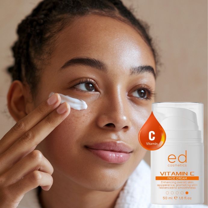 Giovane donna applica crema. Flacone bianco con etichetta arancione. Testo: Vitamin C Face Cream, ed cosmetics.