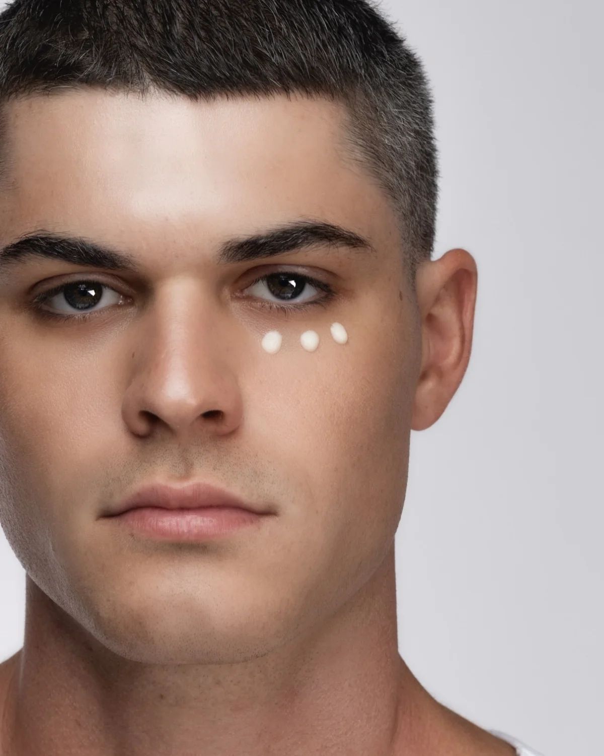 Primo piano di un uomo. Sotto l'occhio sono applicati tre punti di crema chiara.