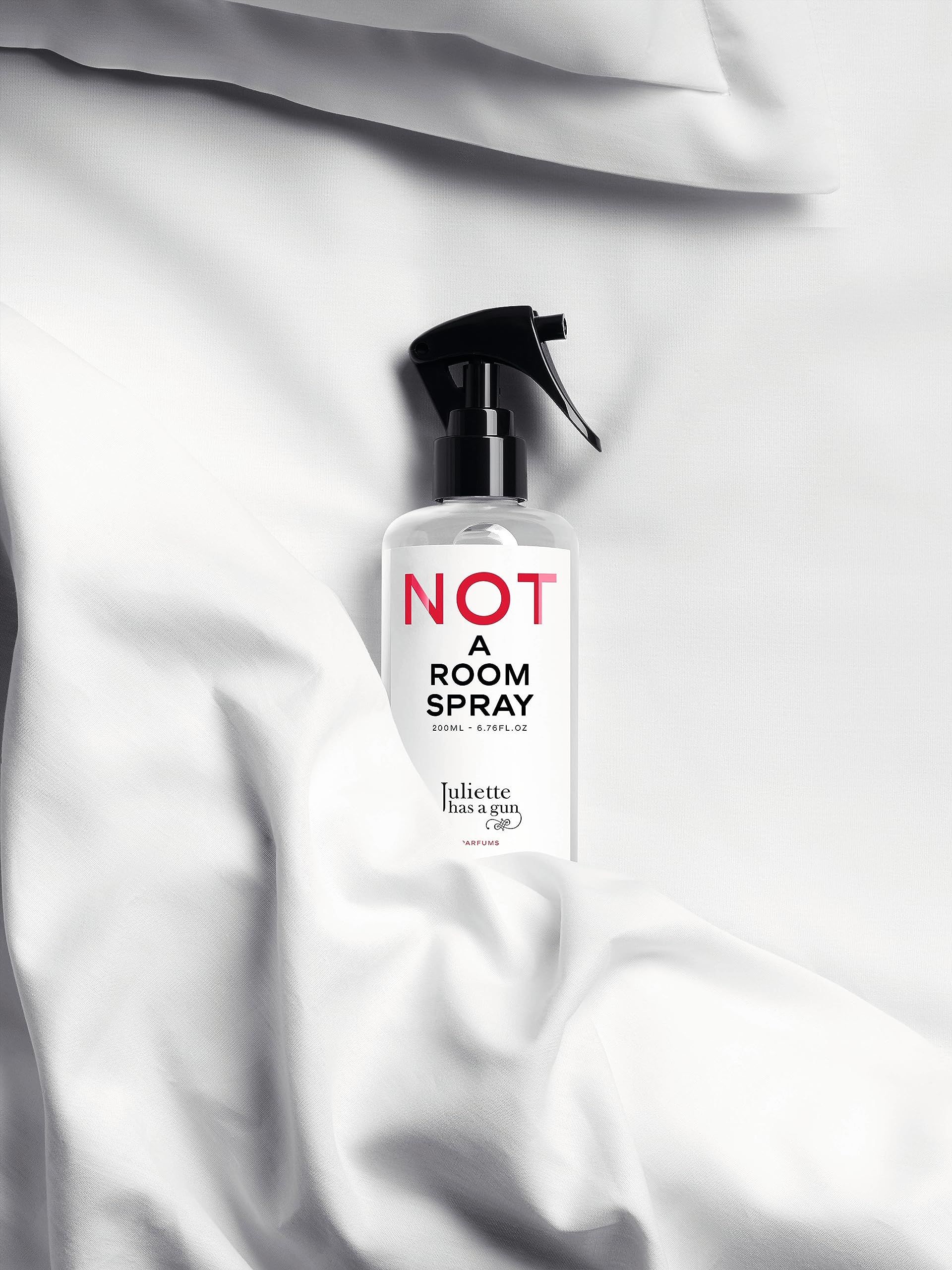 Flacone trasparente con spruzzatore nero su tessuto bianco. Scritta rossa: NOT A ROOM SPRAY. Marchio: Juliette has a gun.