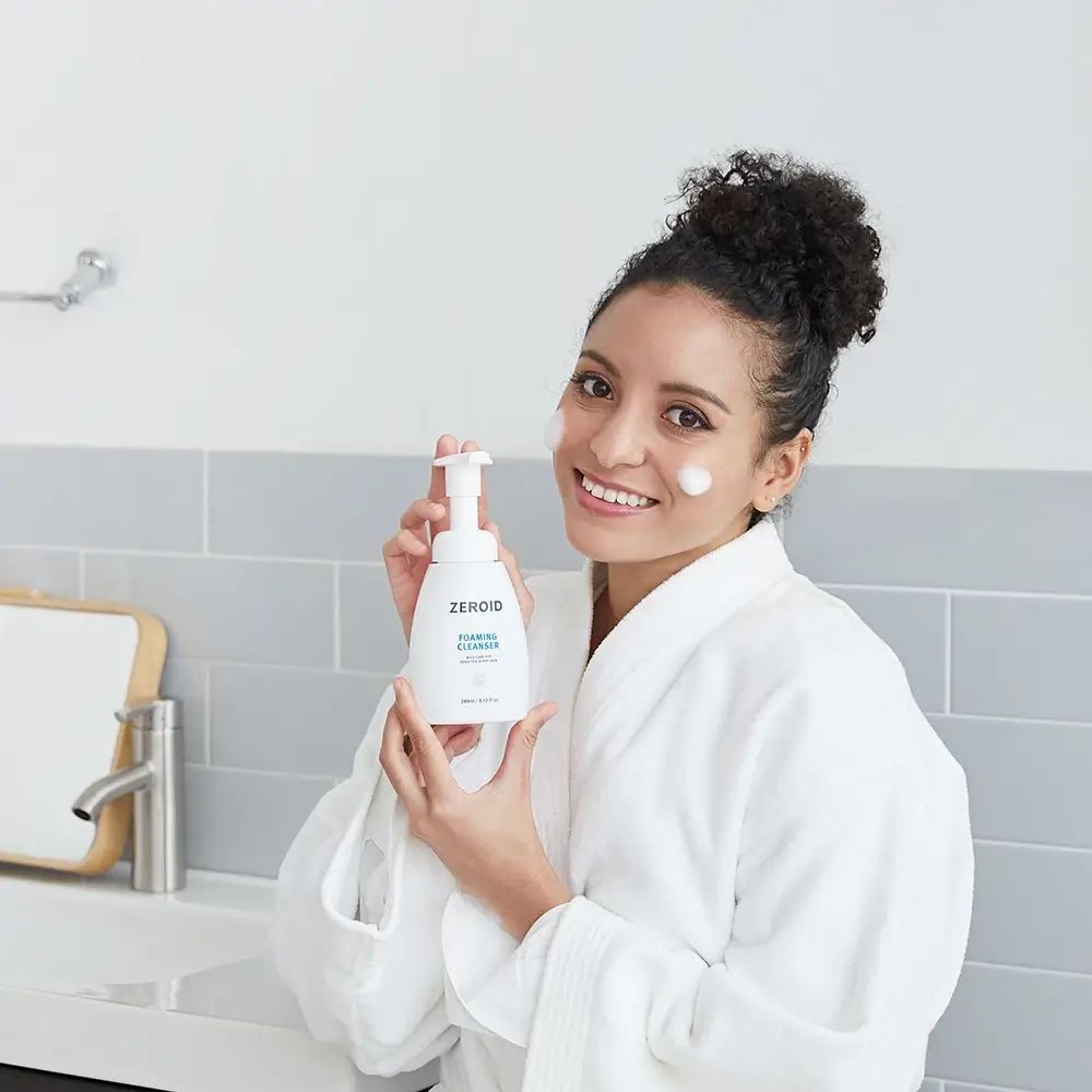 Donna che tiene in mano un flacone bianco ZEROID Foaming Cleanser. Testo: Foaming Cleanser, mild care for sensitive & dry skin. 240ml/8.12 fl oz.