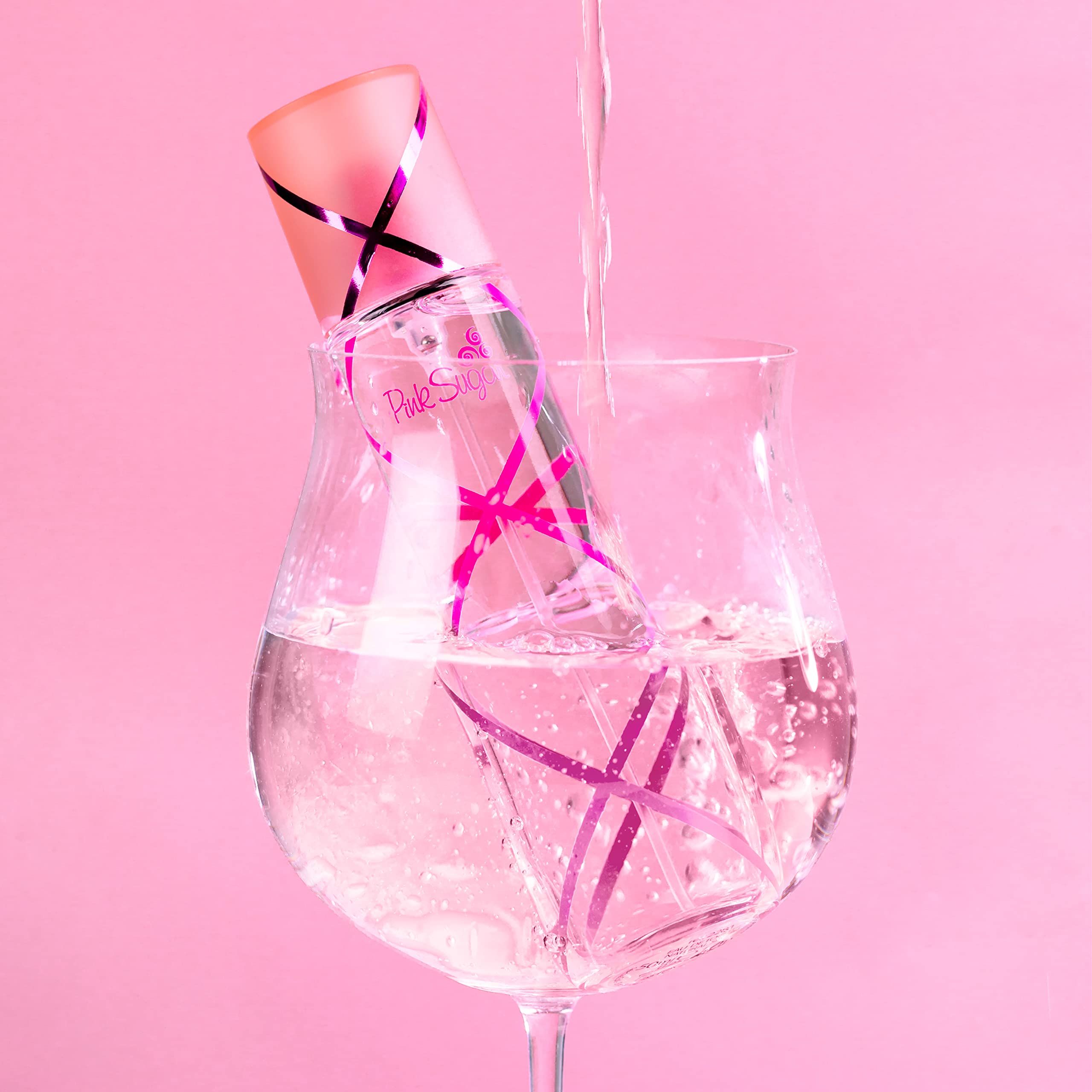 Flacone di profumo in un bicchiere d'acqua. Dettagli rosa e scritta Pink Sugar. L'acqua viene versata nel bicchiere.