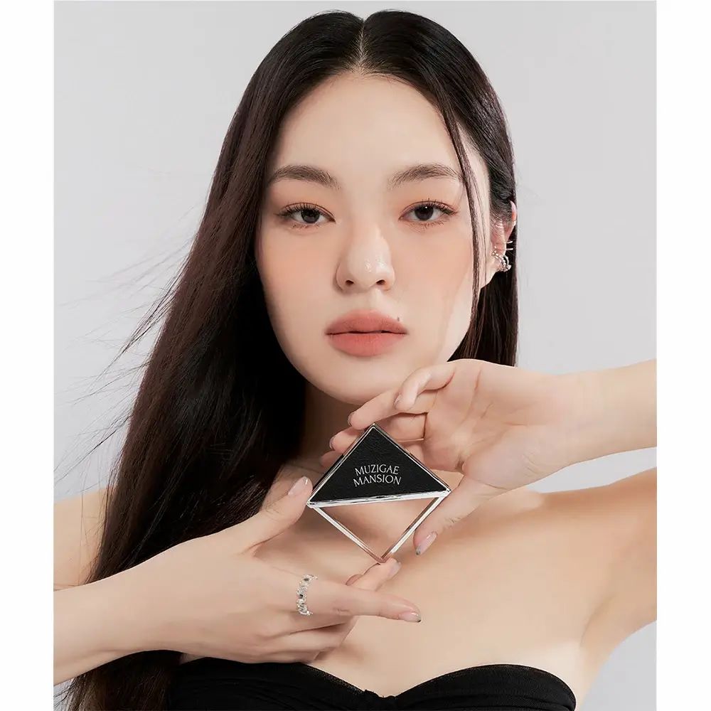 Donna che tiene in mano una palette quadrata MUZIGAE MANSION Fitting Blush. Fard in polvere rosa in un astuccio nero e argento. Donna con fard sulle guance.