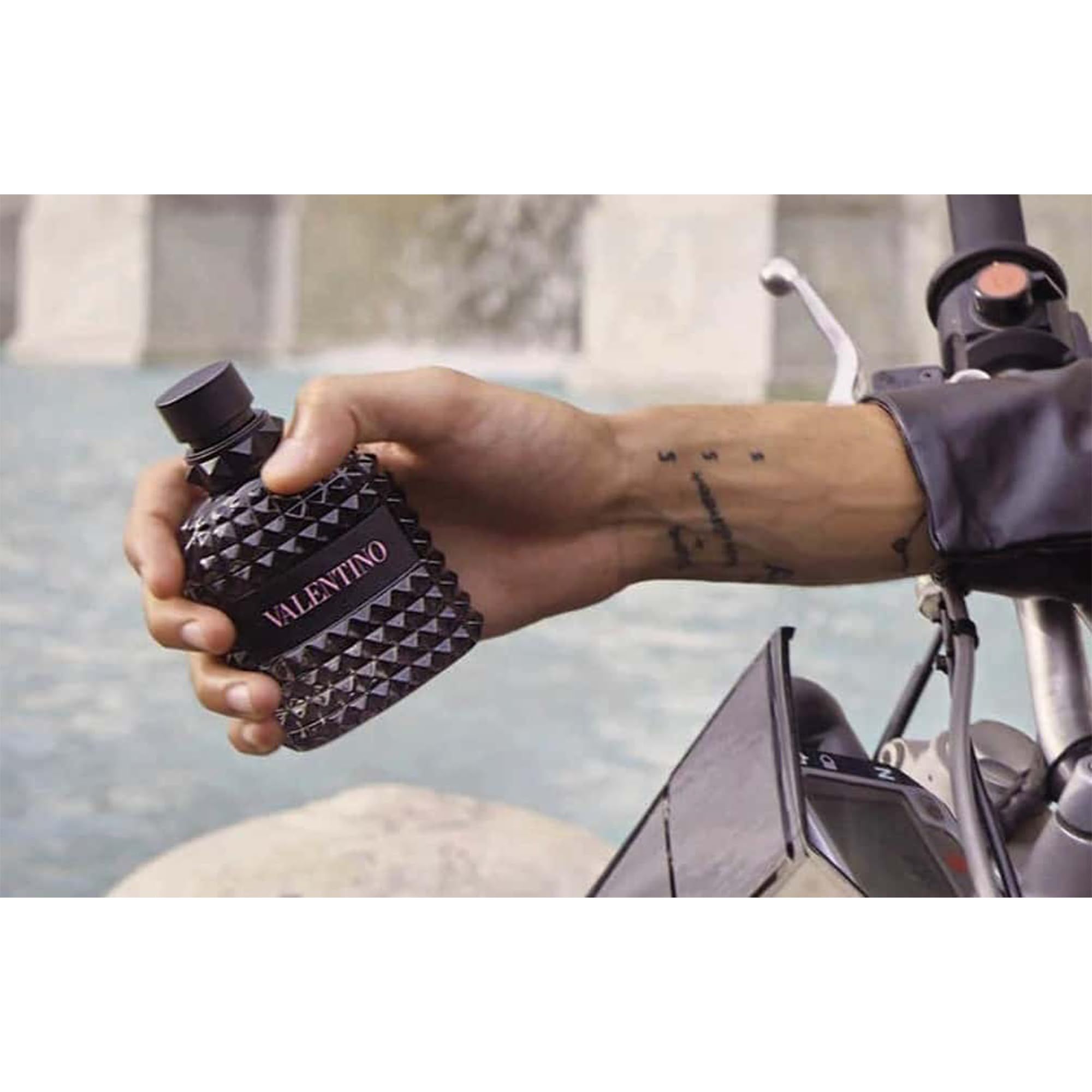 Flacone Valentino tenuto in mano. Tatuaggio sul braccio. Sfondo: moto e acqua.