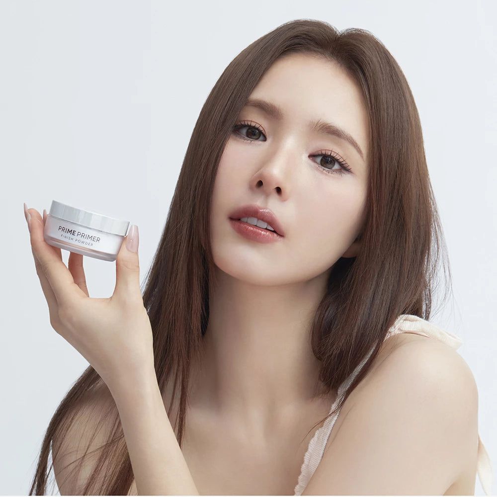 Donna che tiene un vasetto bianco. Scritta: PRIME PRIMER FINISH POWDER. Sorride alla fotocamera.