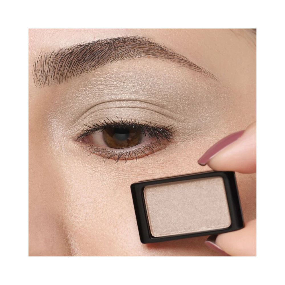 Ombretto in un contenitore nero, tenuto vicino a un occhio truccato. Il colore è chiaro e perlato.