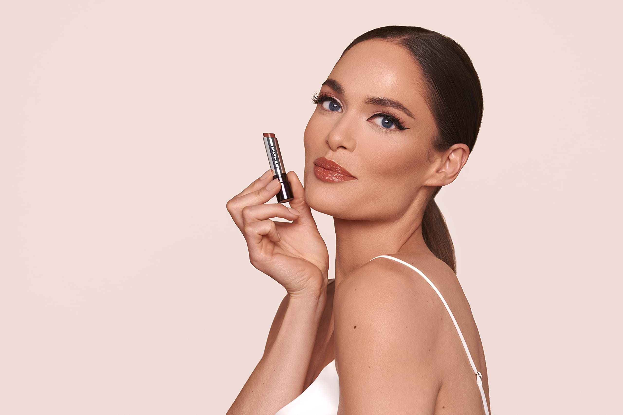 Una donna tiene un balsamo per labbra. Il balsamo per labbra è di By Terry. La donna indossa un top bianco.