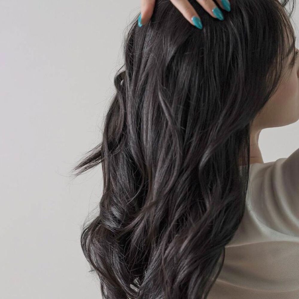 Capelli scuri e mossi, visti da dietro. Una mano con smalto blu tocca i capelli. Lo sfondo è bianco.