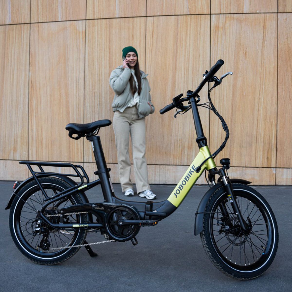 E-bike pieghevole verde-nero. Logo JOBOBIKE. Portapacchi, parafanghi. Persona che telefona vicino alla bici.