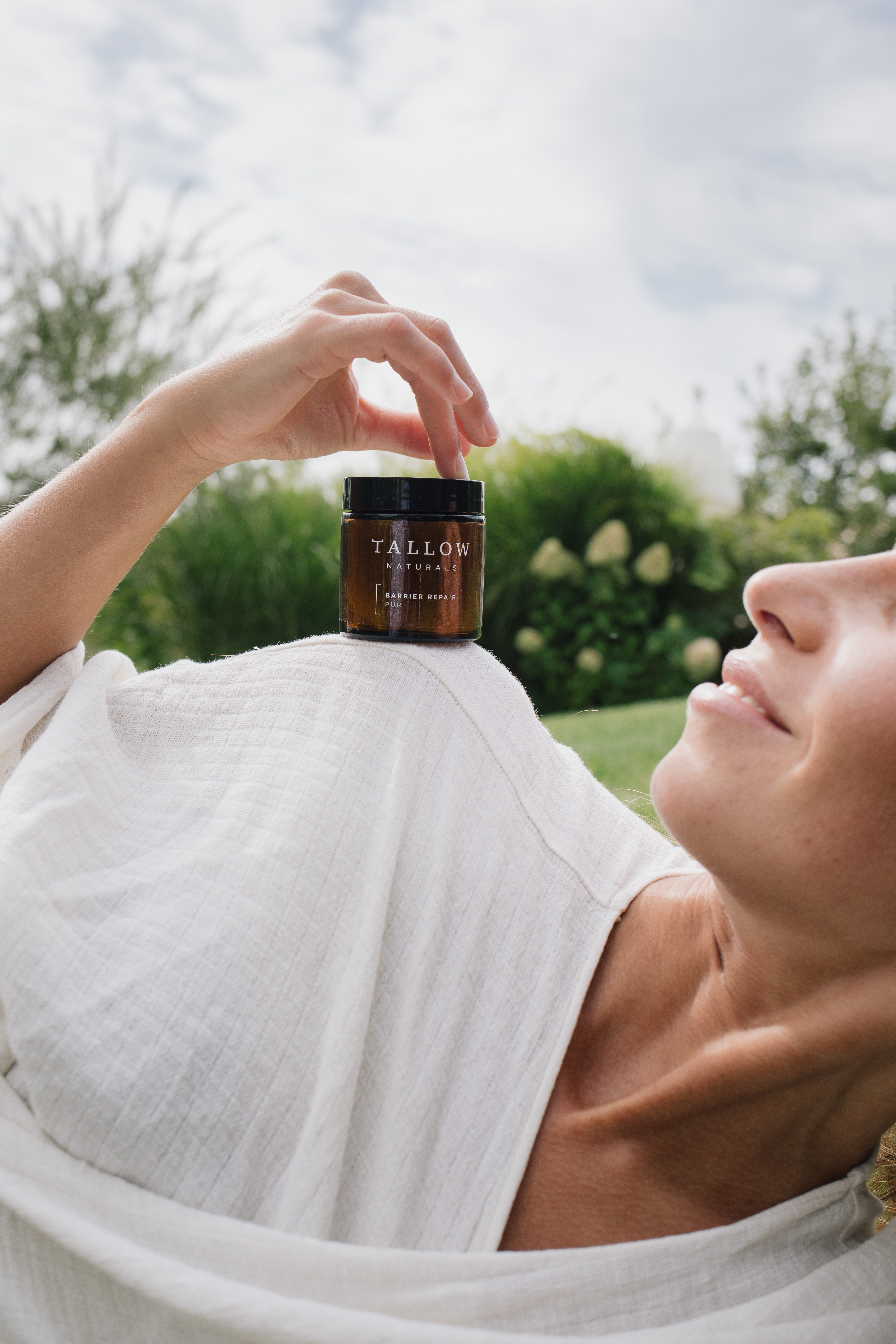 Donna sdraiata all'aperto che tiene un barattolo marrone con coperchio nero. Scritta: TALLOW NATURALS. Nome del prodotto illeggibile.