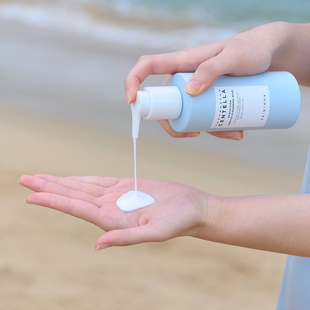 Mani che tengono un flacone azzurro chiaro con erogatore bianco. Il prodotto viene versato nel palmo. Etichetta con testo: SKIN1004.