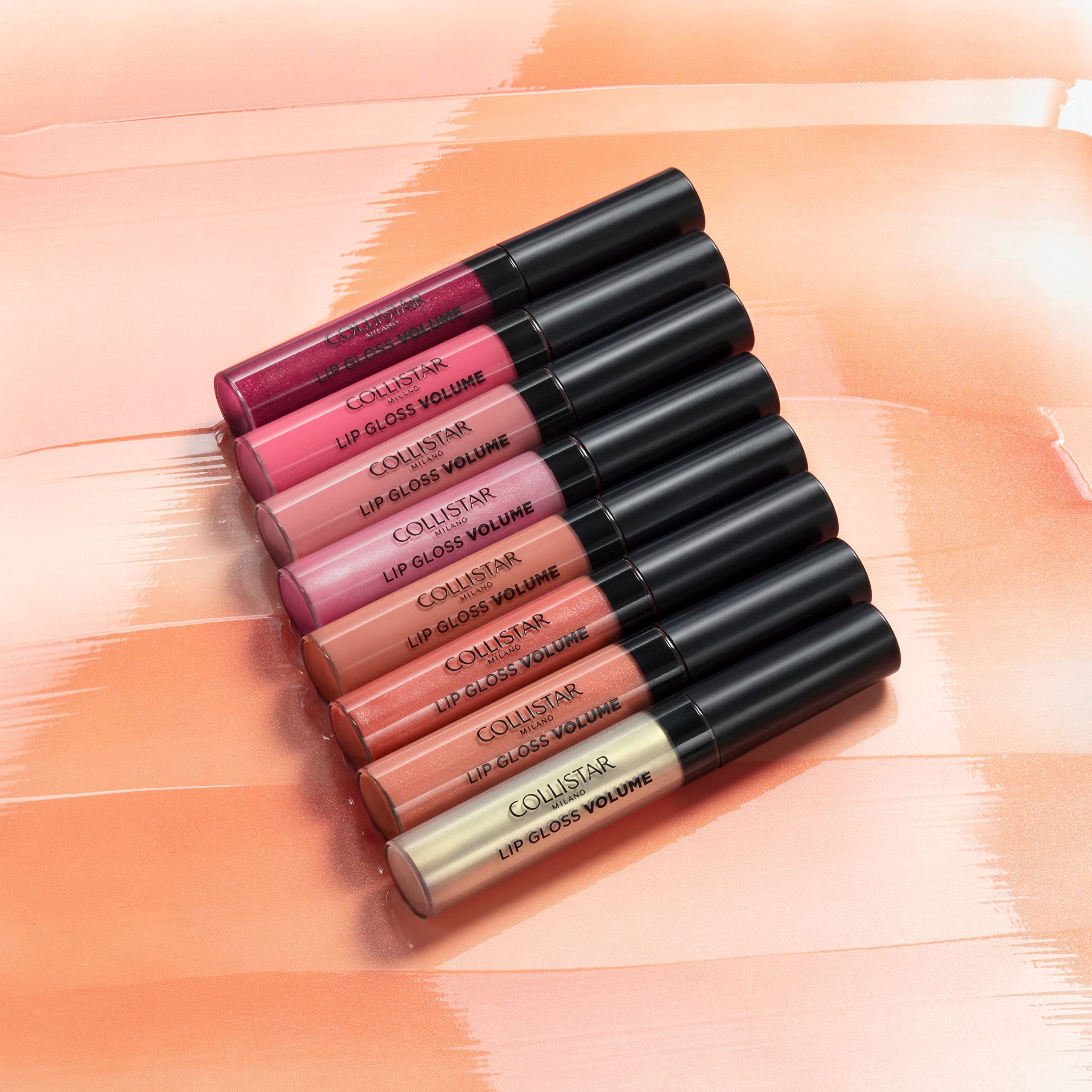 Diversi tubi di lucidalabbra in vari colori, disposti su una superficie colorata. Sui tubi è scritto "COLLISTAR LIP GLOSS VOLUME".