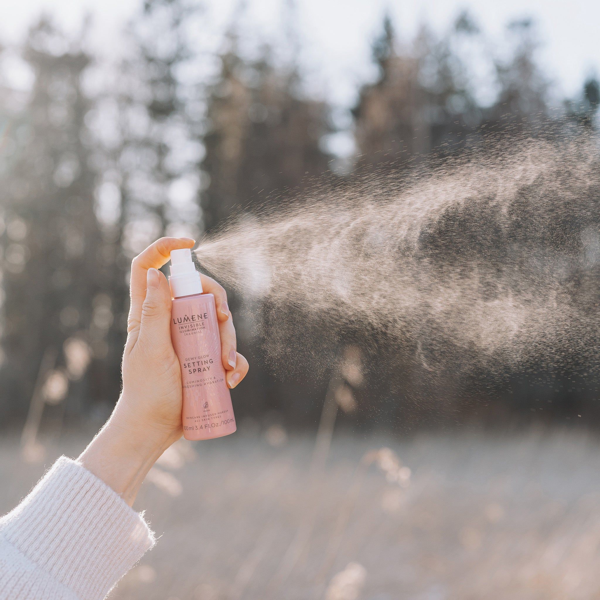 Mano che tiene flacone spray rosa. Nebbia. Scritta: Lumene INVISIBLE ILLUMINATION Dewy Glow Setting Spray. Sfondo: natura, alberi.