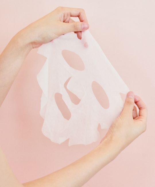 Maschera viso bianca con fori per occhi, naso e bocca, tenuta in mano. Sfondo rosa.