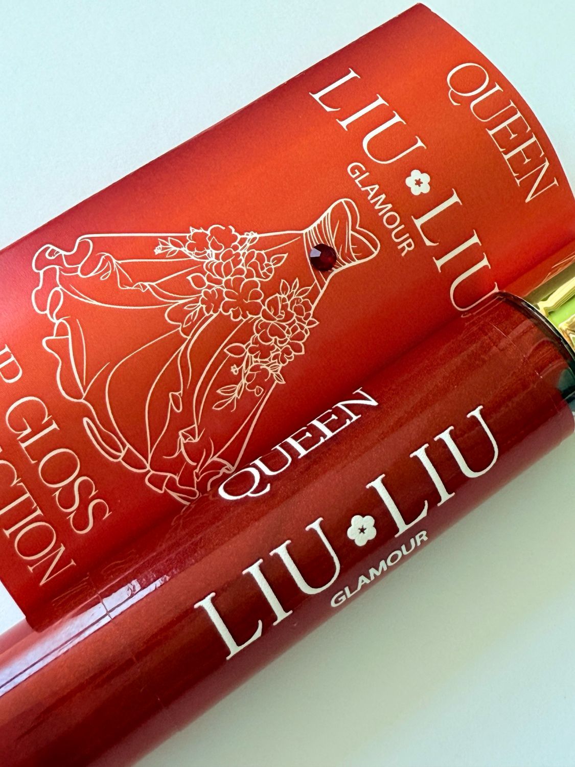 Lucidalabbra rosso e confezione rossa. Scritta: QUEEN LIU LIU Glamour. Confezione con illustrazione di abito da sera. Prodotto e confezione.