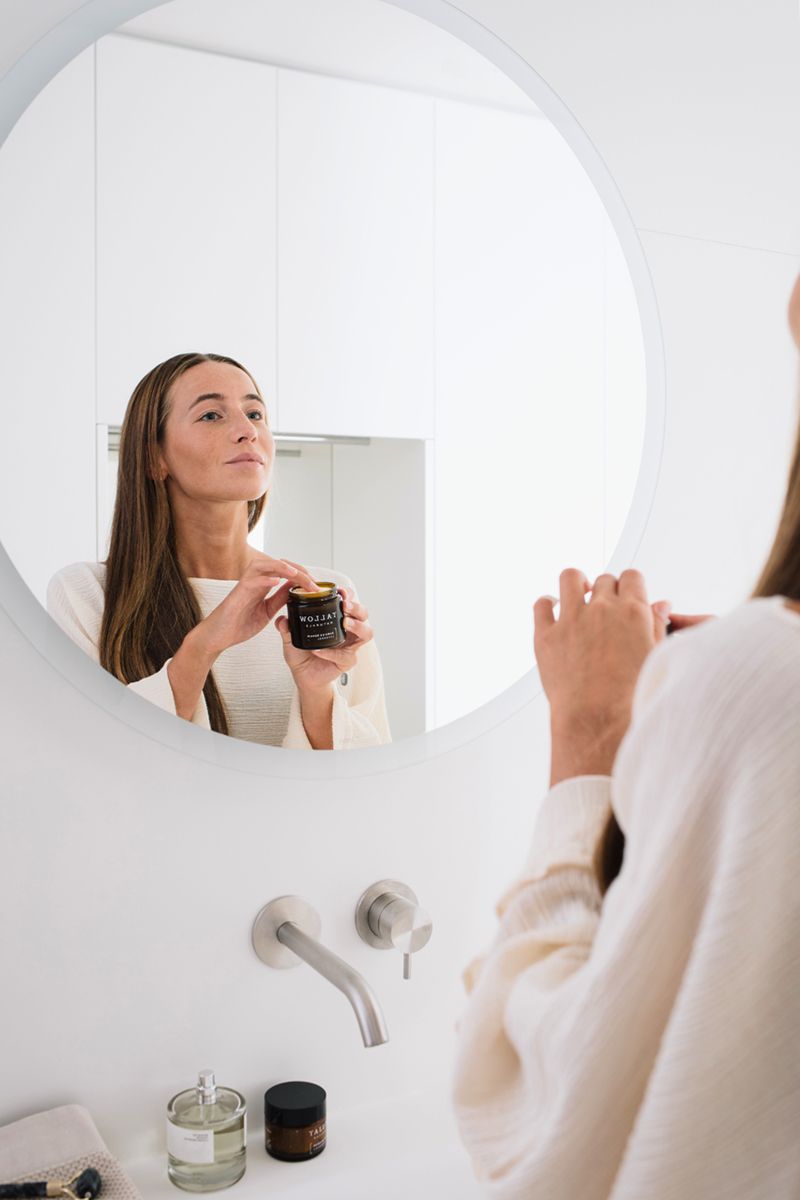 Donna si guarda allo specchio. Tiene un barattolo. Scritta: Tallow Naturals. Scena del bagno.