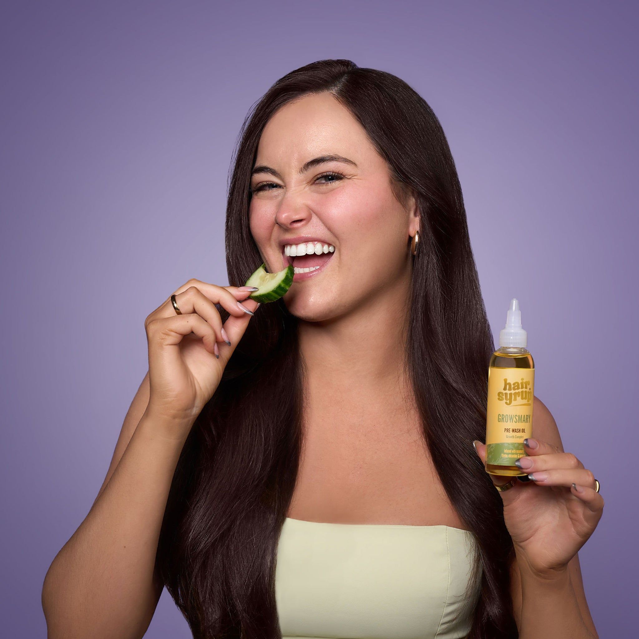 Donna con lunghi capelli scuri mangia cetriolo. Tiene in mano un flacone giallo con etichetta.