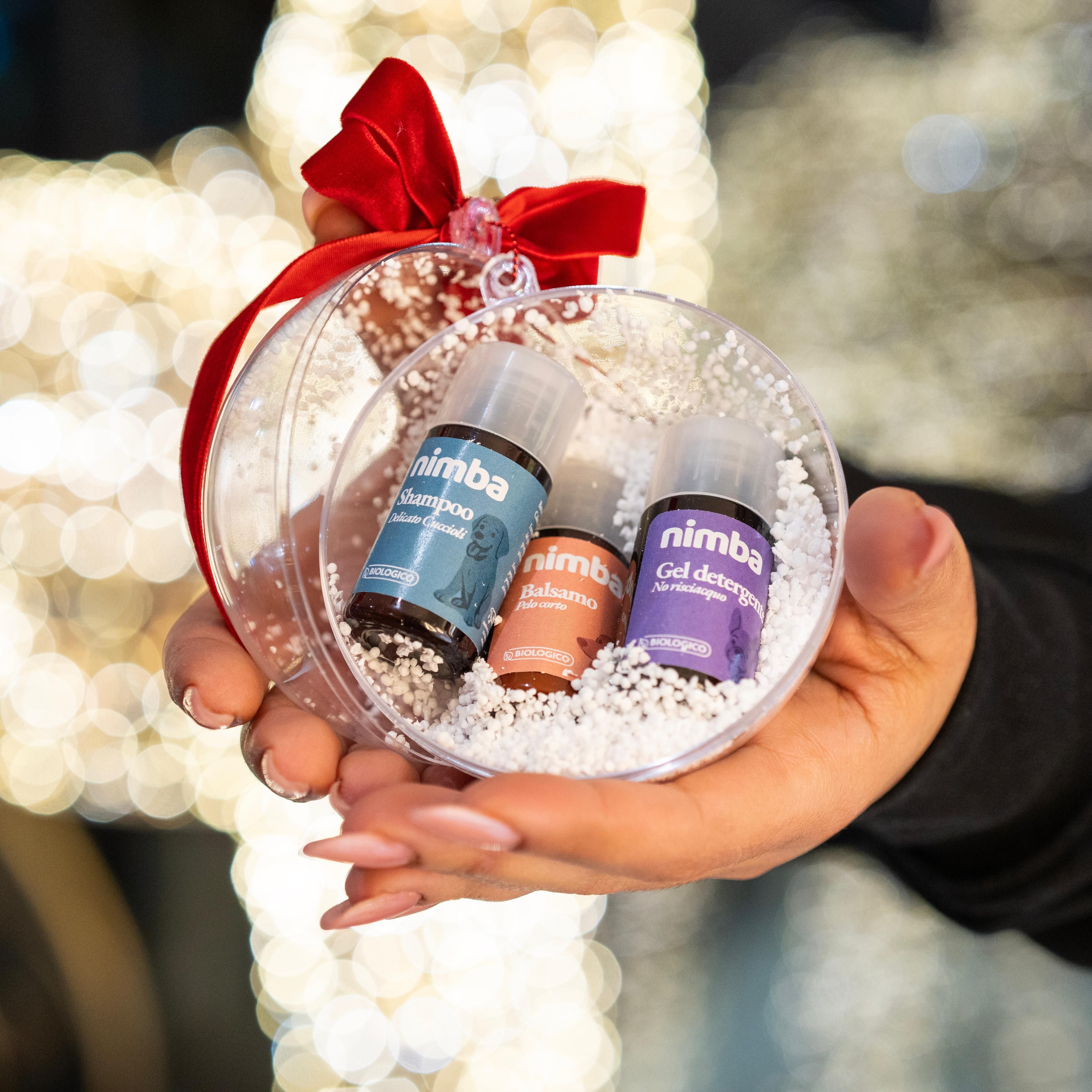 Sfera trasparente natalizia con neve finta e tre flaconi "nimba". Fiocco rosso. Mano che tiene l'ornamento.