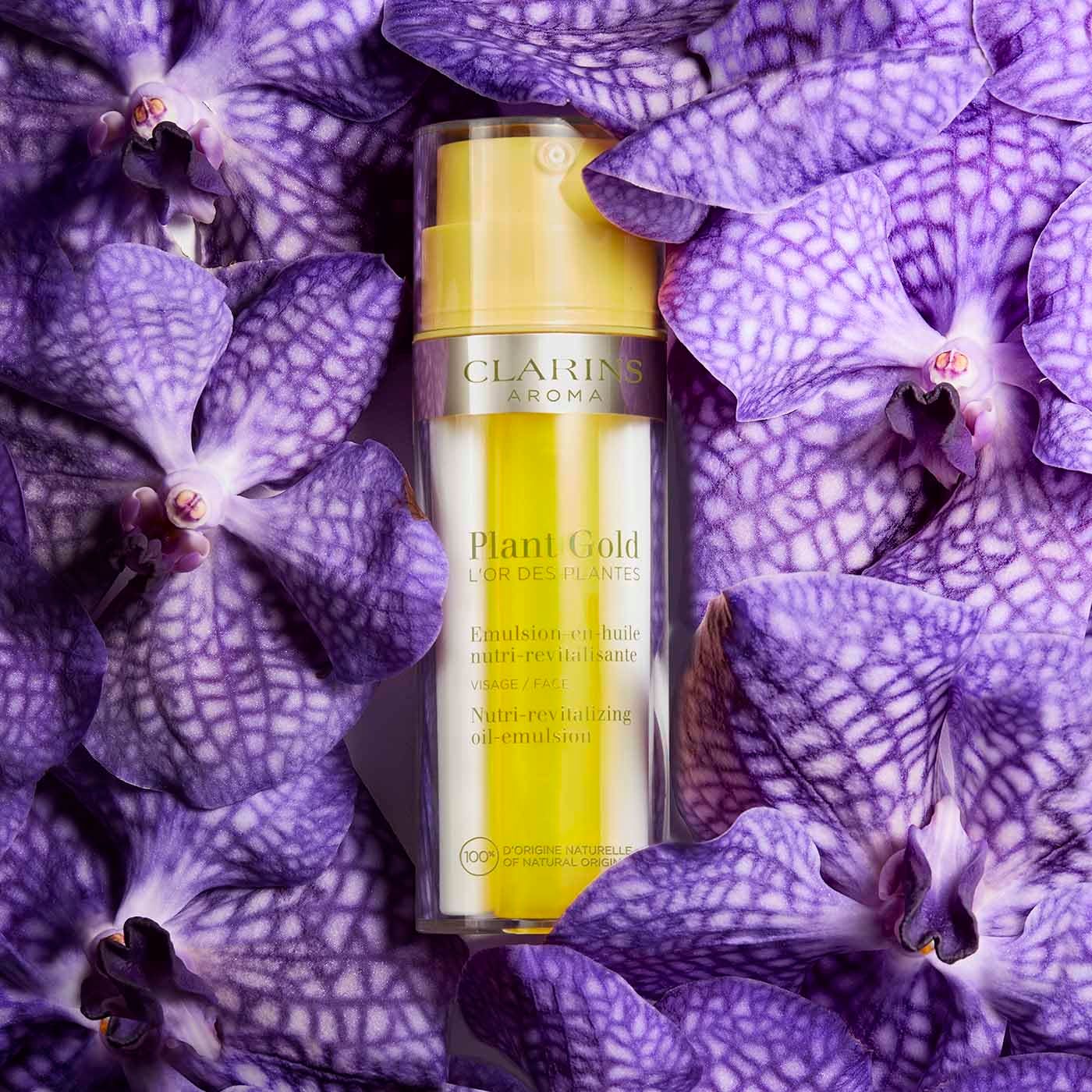 Clarins Plant Gold emulsione olio circondata da orchidee viola. Flacone giallo con pompa, base trasparente. Testo: Emulsion-en-huile.