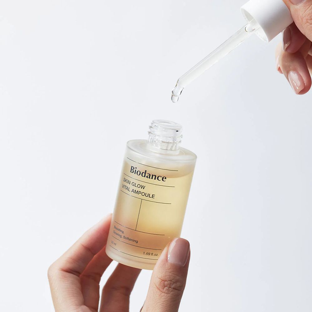 Mani che tengono flacone di siero con pipetta. Gocce cadono dalla pipetta. Scritta: Biodance Skin Glow Vital Ampoule.
