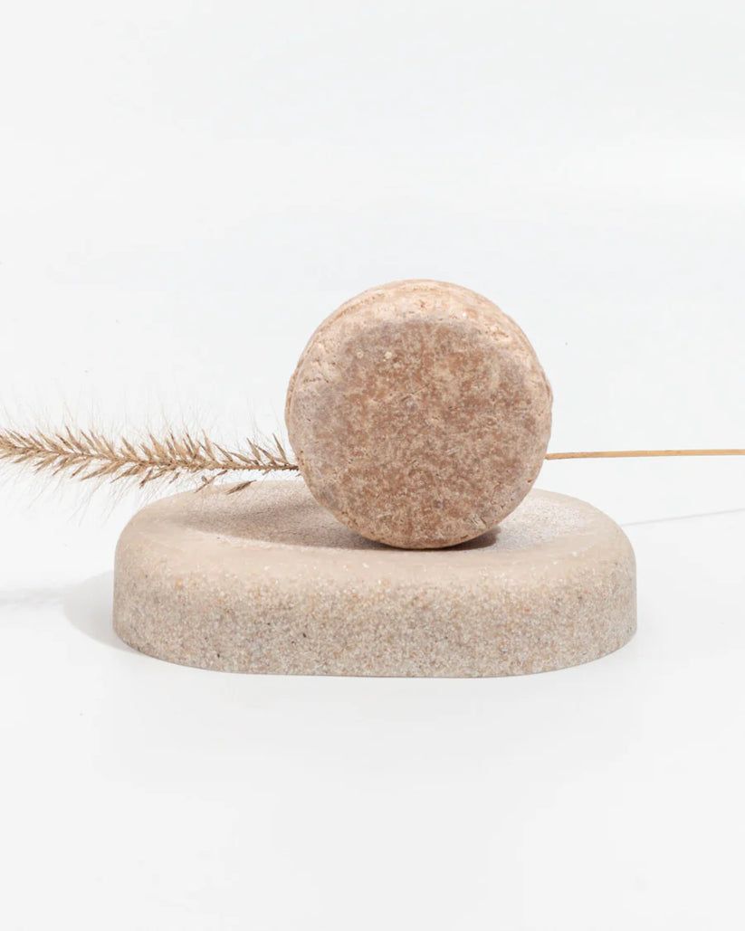 Blocco esfoliante corpo rotondo marrone su un piedistallo abbinato. Erba secca sullo sfondo. Ambiente studio semplice e luminoso.