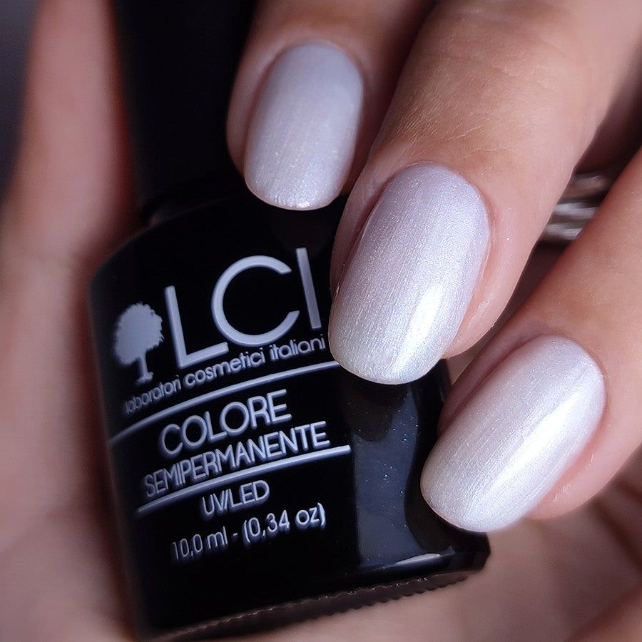 Mano con unghie smaltate e flacone di smalto. Unghie bianco perla. Flacone nero con stampa bianca: LCI, Colore Semipermanente.