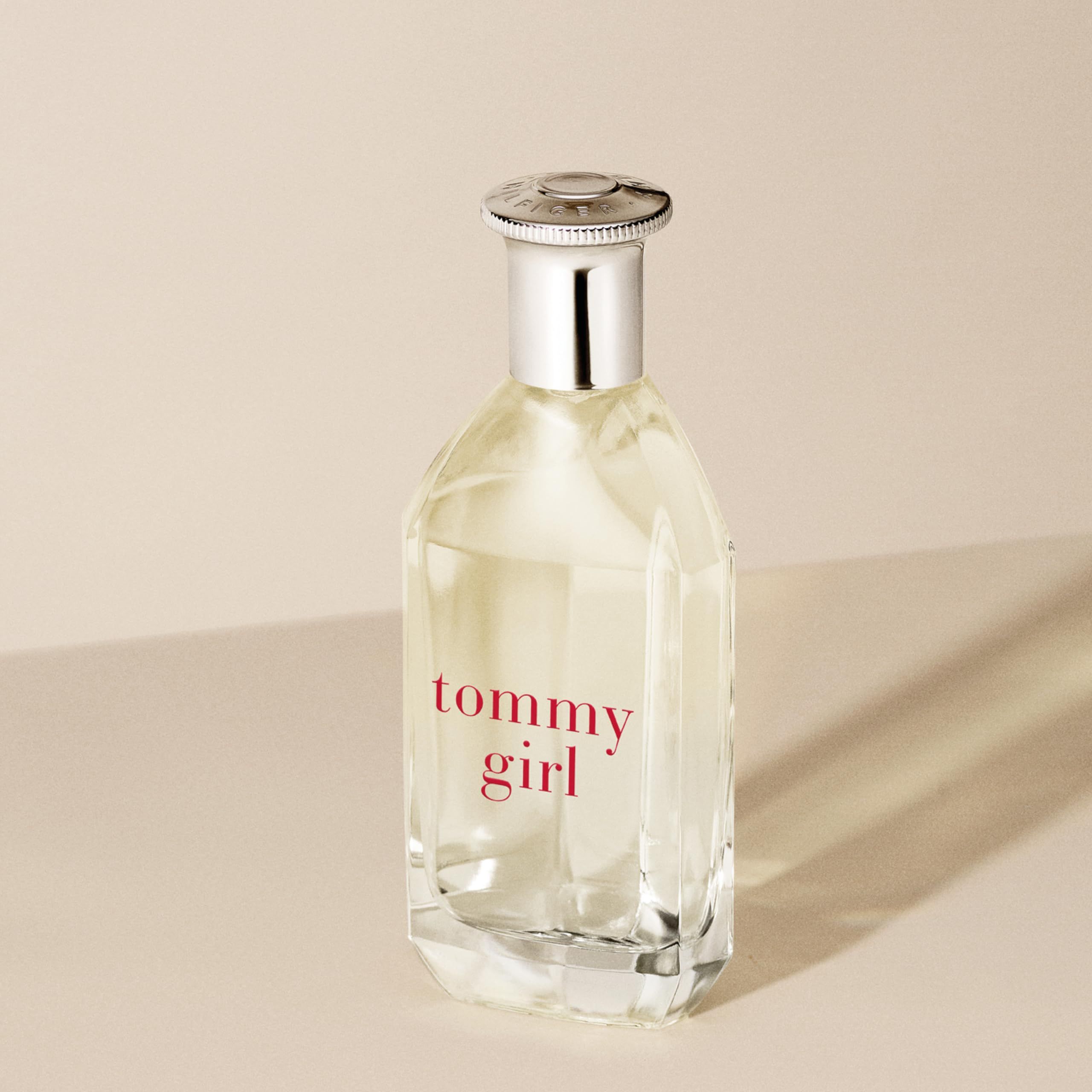 Flacone di profumo con tappo argentato e scritta rossa "tommy girl". Flacone trasparente, rettangolare, su sfondo beige.