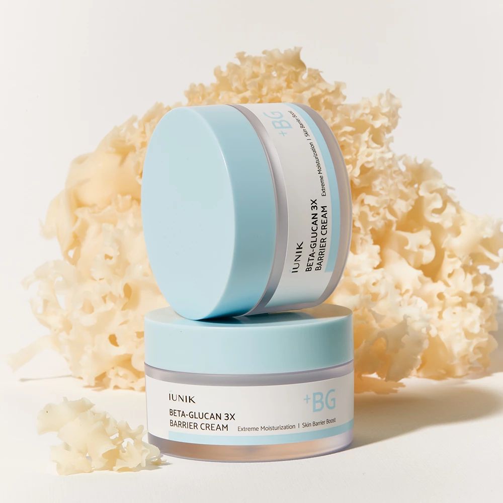 Due barattoli di crema con coperchi azzurri. Scritta: iUNIK Beta-Glucan 3X Barrier Cream. Sfondo: tessuto bianco floreale.