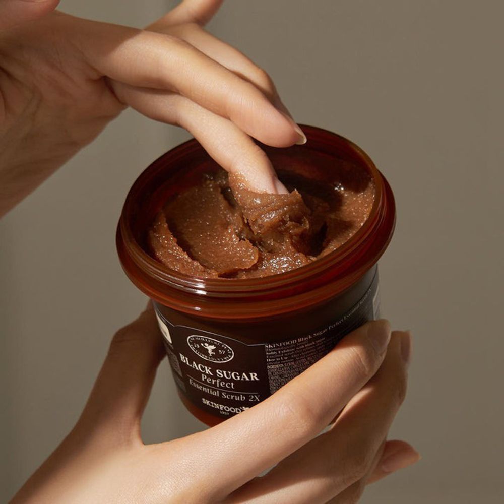 Mani che tengono un vasetto marrone aperto. Dita che toccano lo scrub. Scritta: Black Sugar Perfect Essential Scrub 2X.