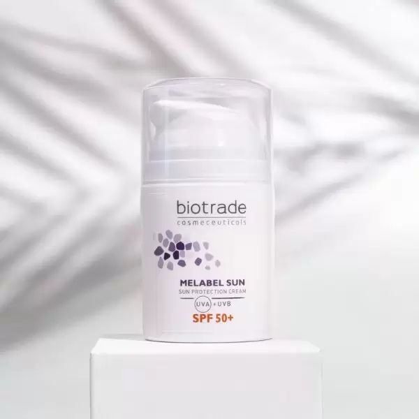 Flacone bianco Biotrade Melabel Sun SPF 50. Scritta: Melabel Sun, SPF 50+. Su piedistallo bianco. Ombre di foglie sullo sfondo.