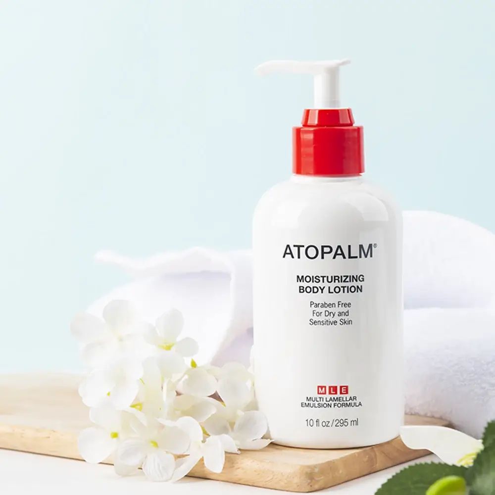 Flacone bianco con erogatore rosso, accanto a fiori e asciugamano. Scritta: ATOPALM Moisturizing Body Lotion.