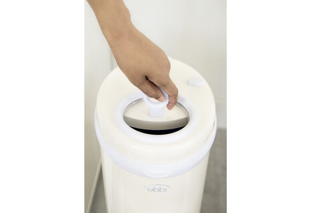 Primo piano del coperchio di un bidone per pannolini beige. Mano che apre il coperchio bianco. Marchio 'ubbi'.