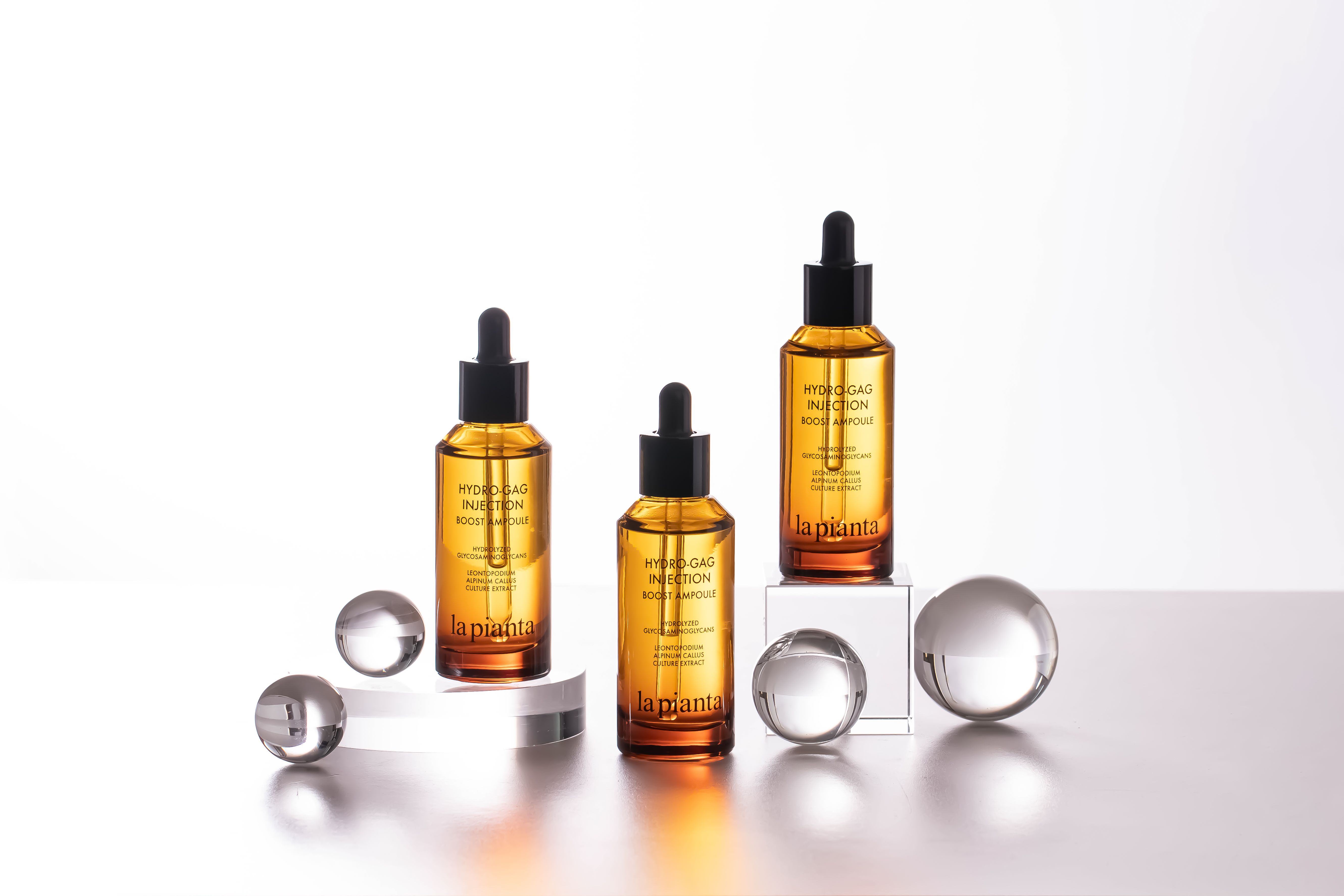 Tre flaconi di vetro marrone con contagocce nero. Su piedistallo e con sfere. Scritta: HYDRO-GAG INJECTION BOOST AMPOULE.