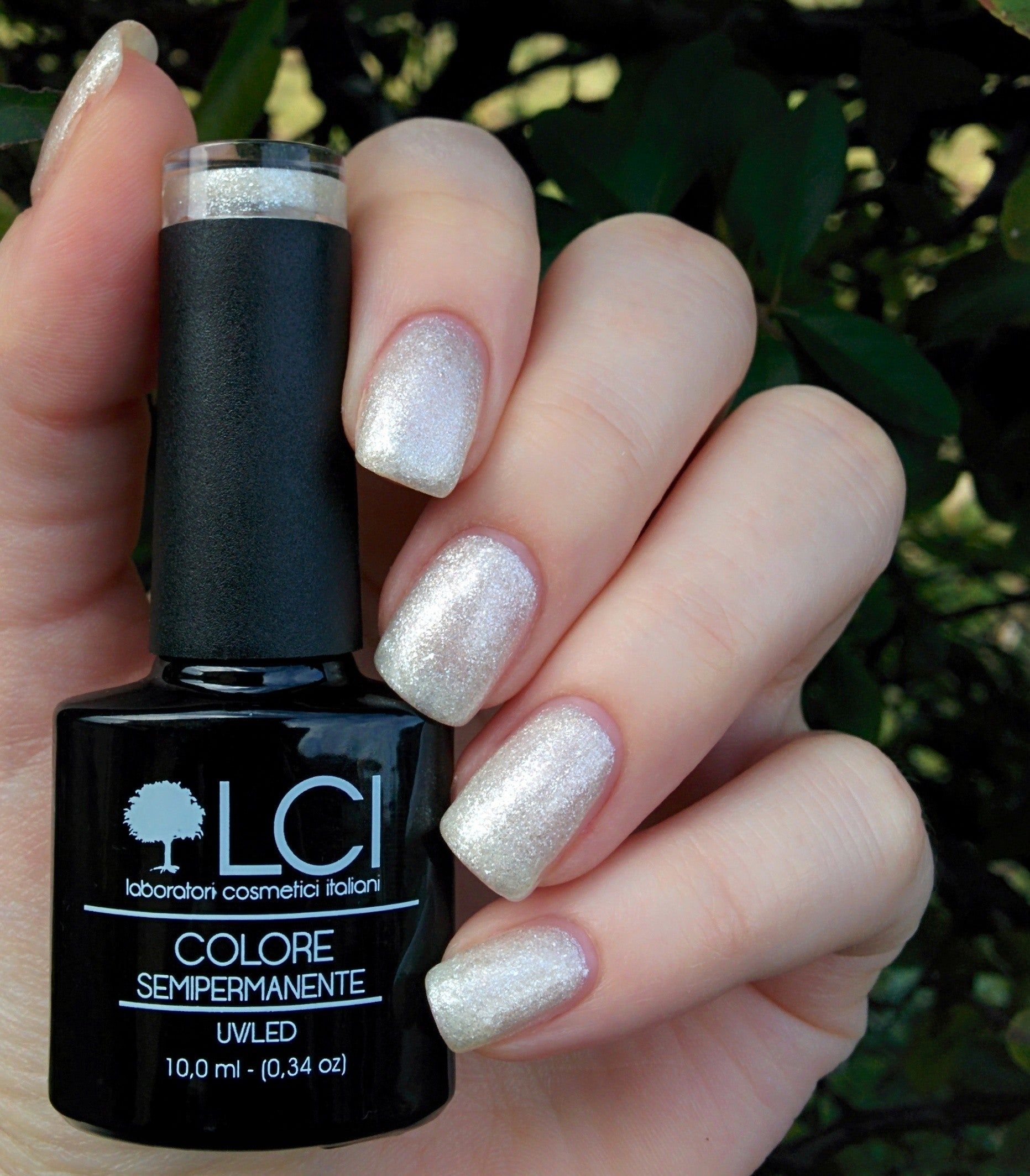 Flacone di smalto per unghie accanto a unghie smaltate. Scritta: LCI, Colore Semipermanente, UVLED 10,0 ml. Unghie con smalto bianco glitterato.