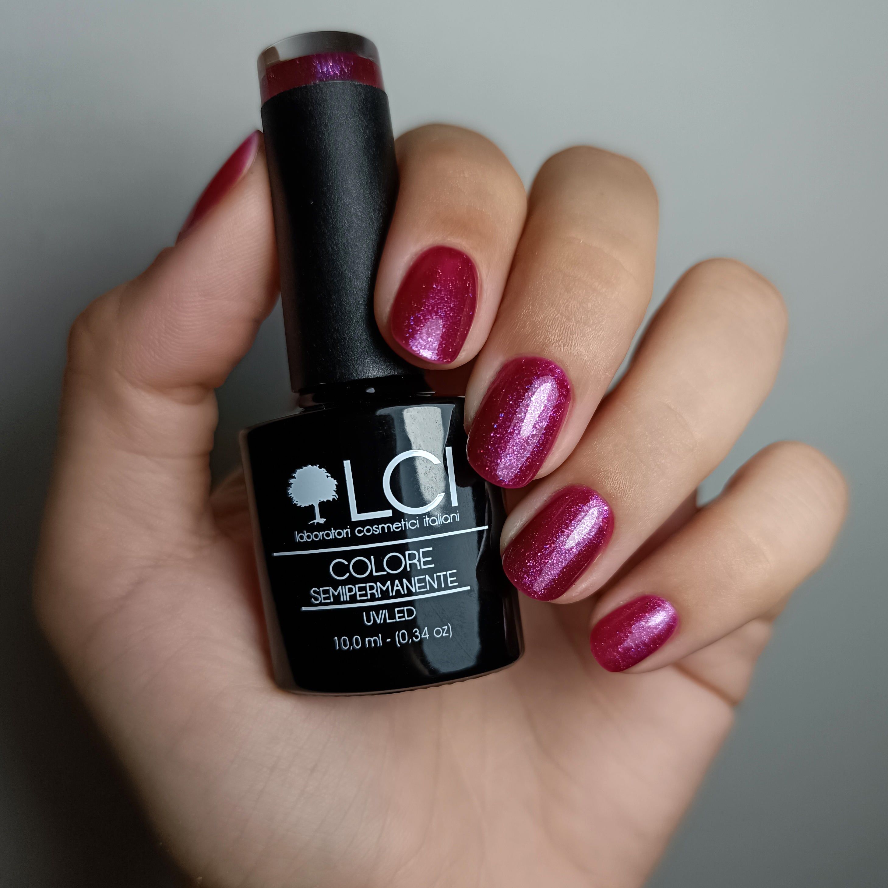 Mano con unghie smaltate e flacone di smalto. Colore: viola con glitter. Scritta: LCI, Colore Semipermanente, UVLED.