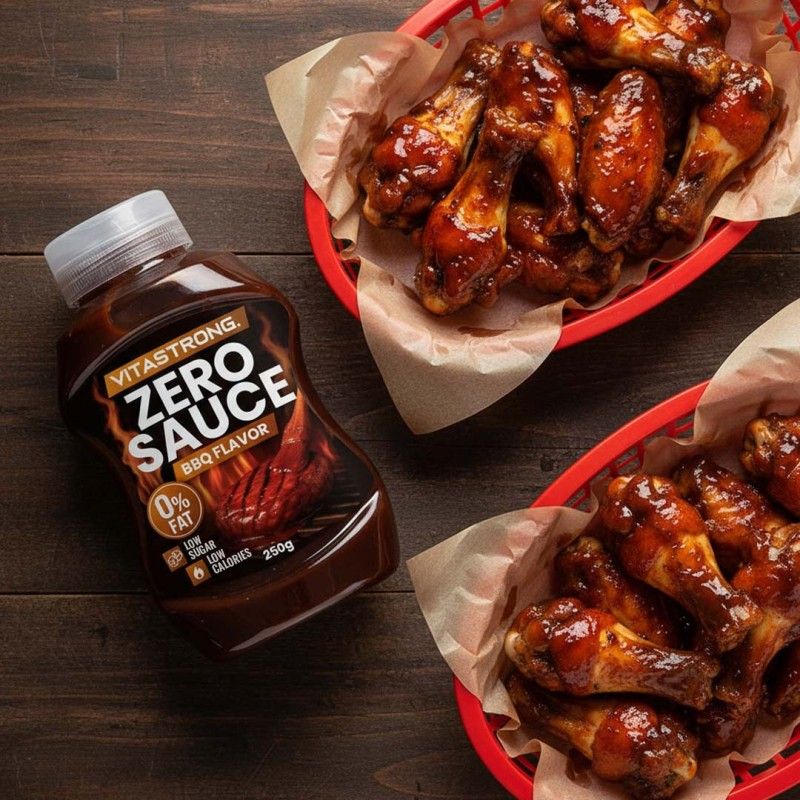 Bottiglia marrone con tappo bianco. Scritta: Vitastrong Zero Sauce BBQ Flavor. 0% grassi, basso contenuto di zuccheri, basse calorie. 250g. Accanto a due cestini di ali di pollo.