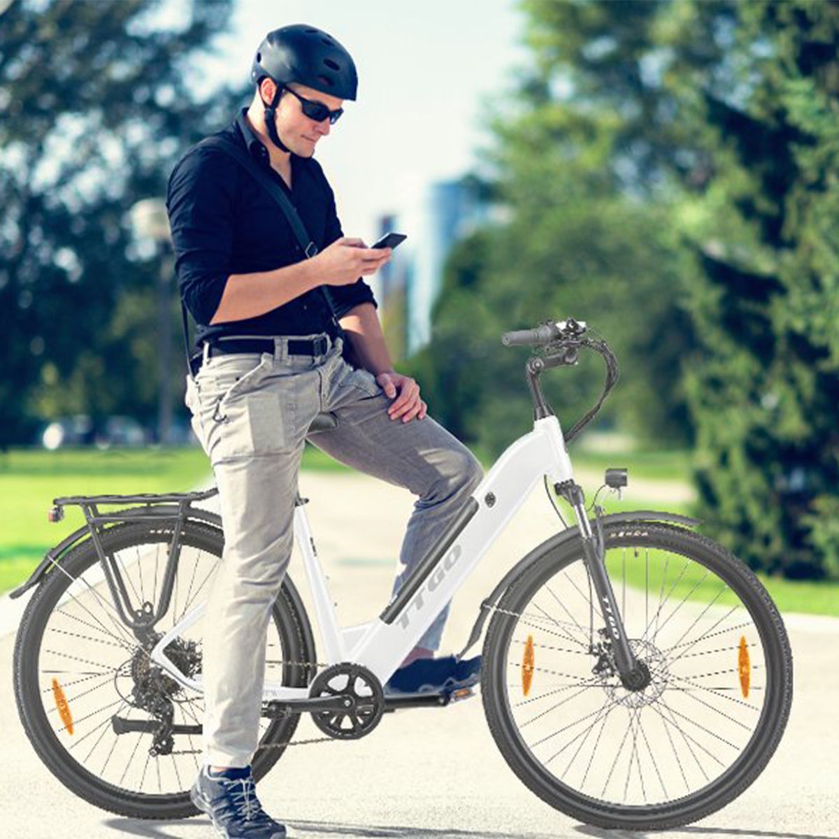 Uomo in piedi accanto a e-bike bianca. Casco, occhiali da sole e smartphone. Sfondo: alberi ed edifici.