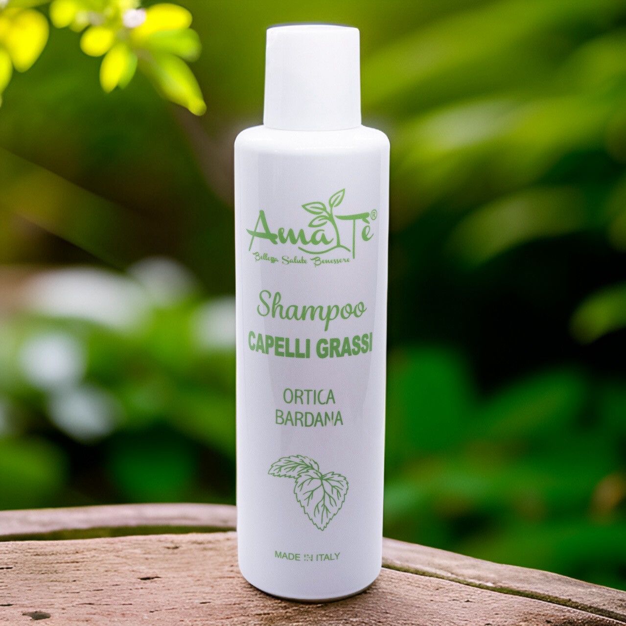 Flacone bianco con etichetta verde. Testo: Amatè Shampoo Capelli Grassi Ortica Bardana. Sfondo: fogliame verde.