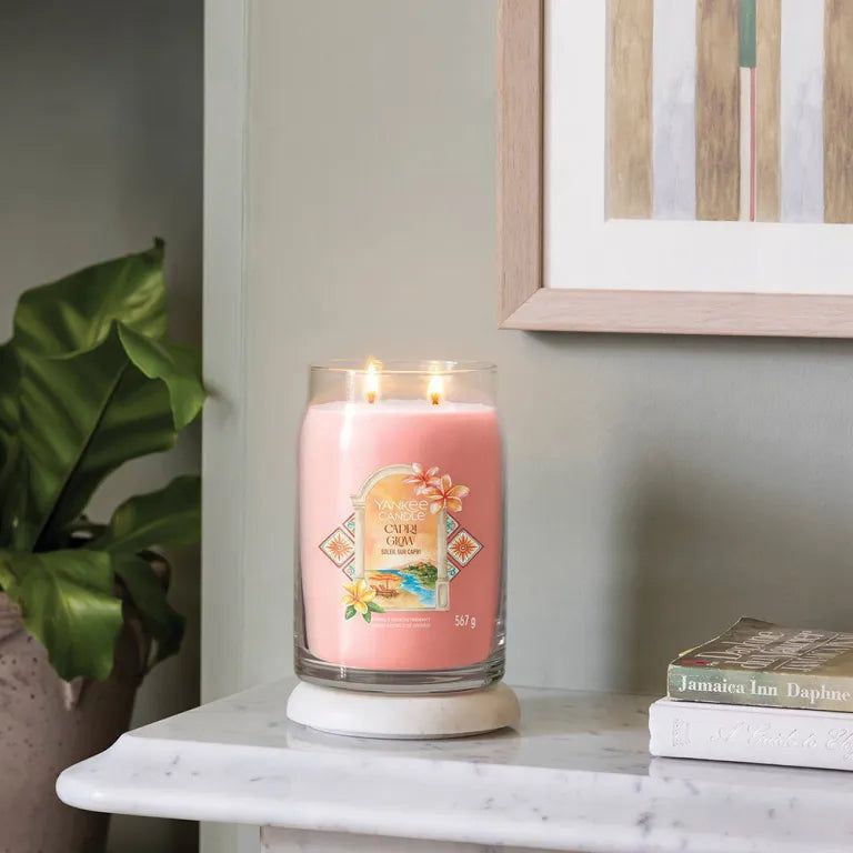 Candela rosa in vetro, due stoppini, su base bianca. Scritta: Capri Glow. Elementi decorativi.