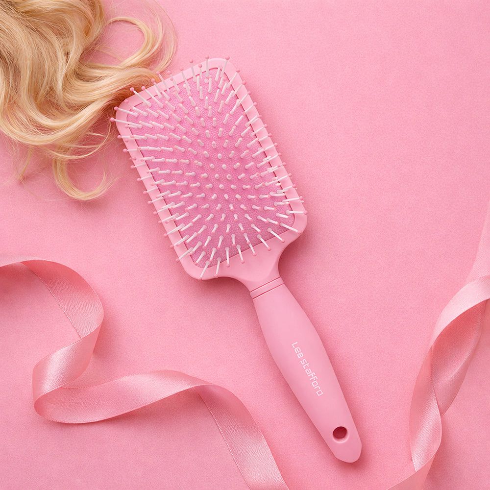 Spazzola piatta rosa con setole bianche. Il manico riporta il nome del marchio. Capelli biondi e nastro rosa sullo sfondo.