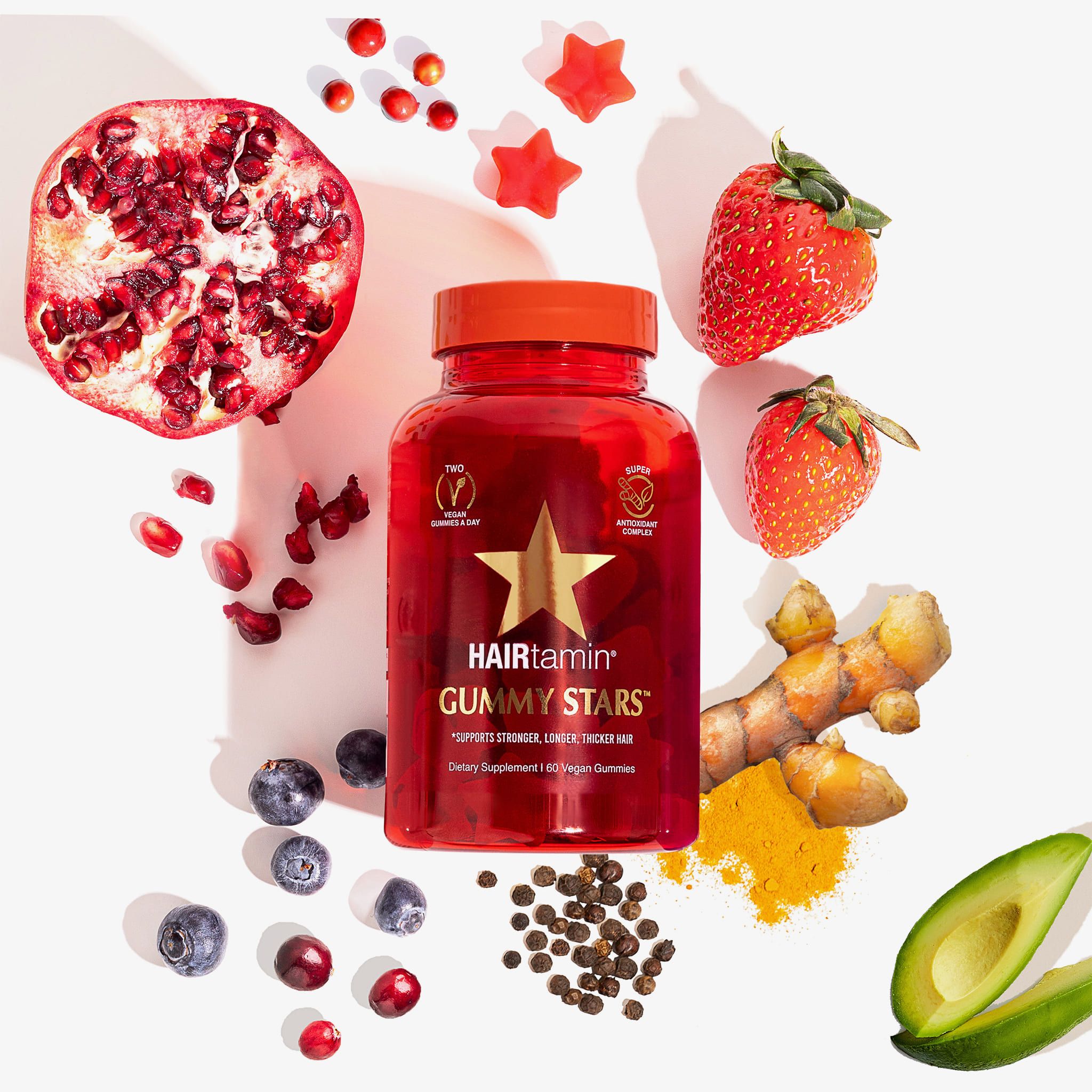 Bottiglia rossa HAIRtamin Gummy Stars, circondata da frutta e spezie: melograno, fragole, mirtilli, curcuma.