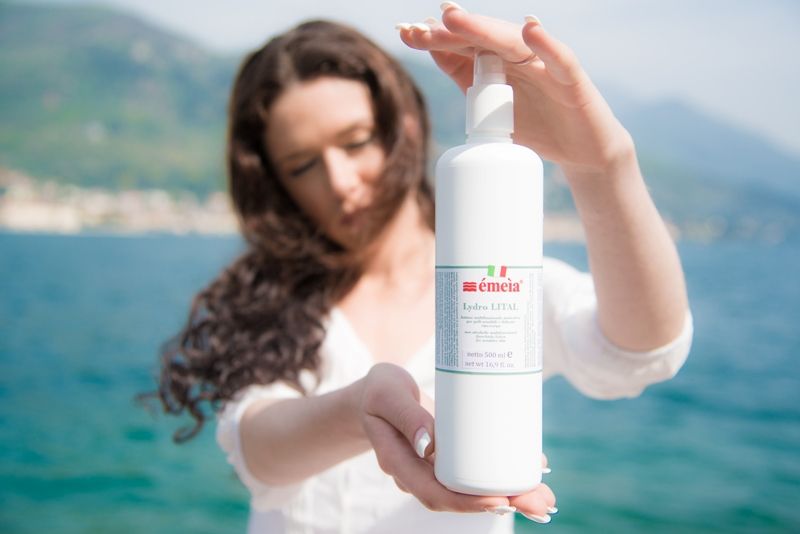 Donna tiene flacone spray bianco Chémeìa Lydro-LITAL con etichetta, mare e cielo sullo sfondo.