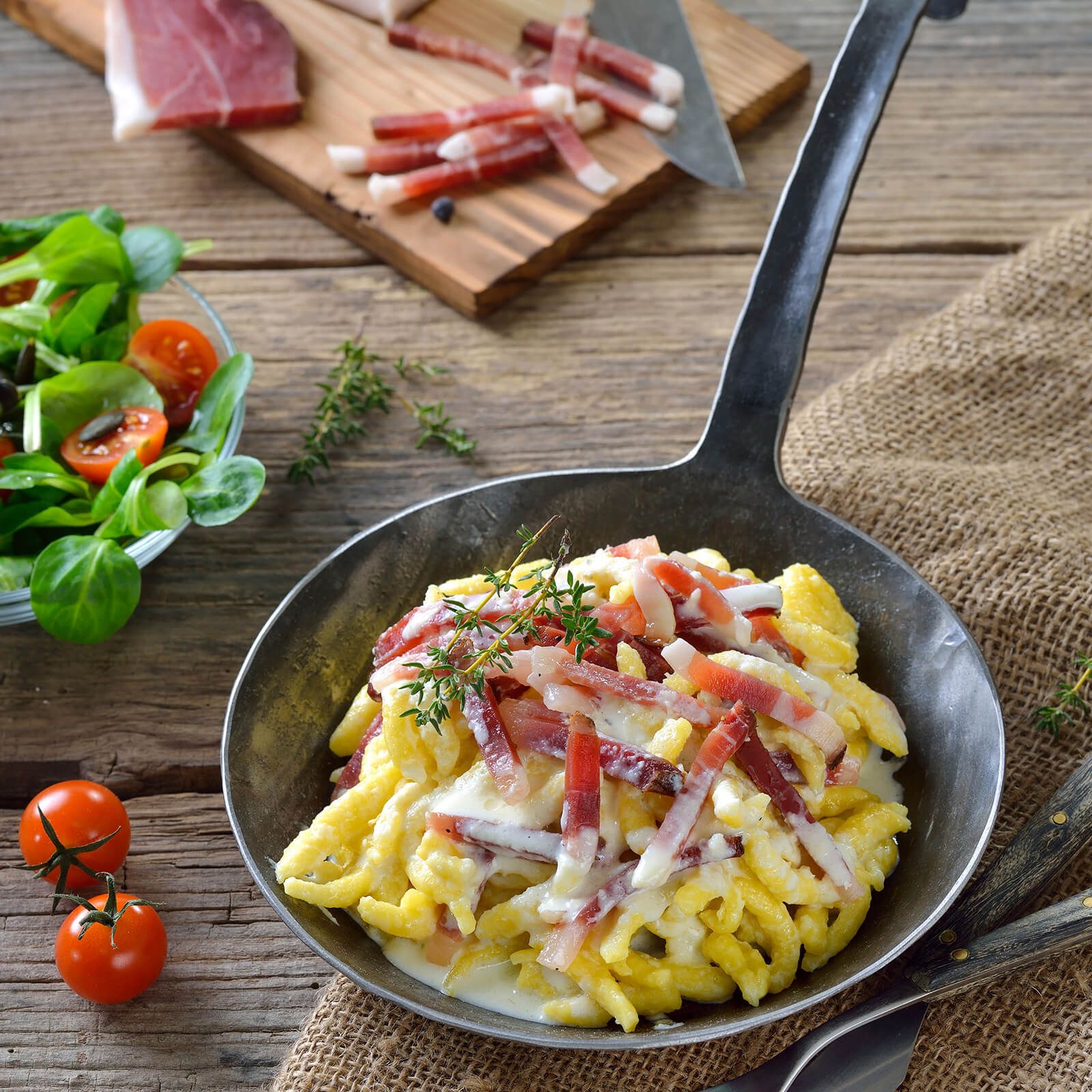 Padella con spätzle, pancetta e salsa. Su un tavolo di legno con ingredienti e decorazioni.